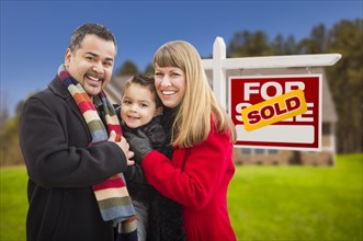 Warmly dressed young mixed-race family in front of sold home for sale real estate sign and house
