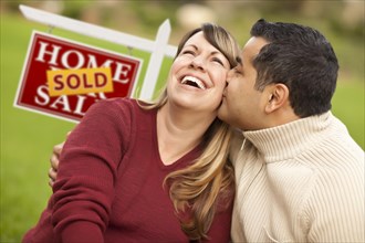 Happy mixed-race couple in front of sold real estate sign