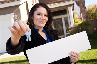 Happy attractive hispanic woman holding blank sign and keys in front of house