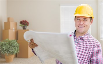 Happy male construction worker in room with moving boxes holding roll of blueprints