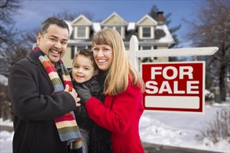 Warmly dressed young mixed-race family in front of home for sale real estate sign and house with snow on the ground