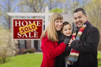Warmly dressed young mixed-race family in front of sold home for sale real estate sign and house
