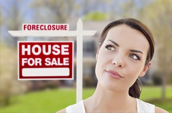 Thoughtful pretty mixed-race woman in front of home and foreclosure house for sale real estate sign looking up and to the side
