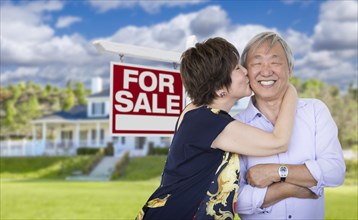 Attractive affectionate senior chinese couple in front of beautiful house and for sale real estate sign