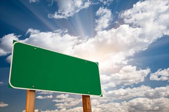 Blank green road sign over dramatic blue sky with clouds and sunburst