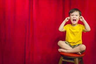mixed-race boy with his fingers in his ears sitting on stool in front of red curtain