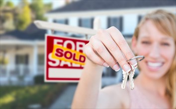 Excited woman holding house keys and sold for sale real estate sign in front of nice new home