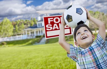 Playful young boy holding soccer ball in front of house and for sale real estate sign