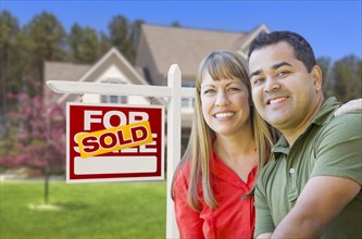 Happy mixed-race couple in front of sold home for sale real estate sign and house