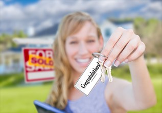 Woman holding new house keys with congratulations card in front of sold real estate sign and home