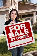 Happy attractive hispanic woman holding for sale by owner real estate sign in front of house