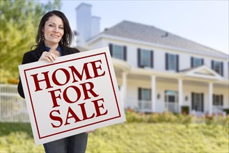 Smiling hispanic female holding sold for sale sign in front of beautiful house