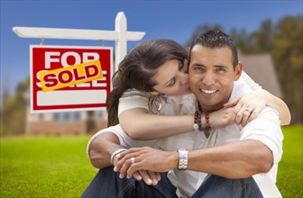 Young happy hispanic young couple in front of their new home and sold for sale real estate sign