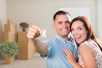 Happy young military couple with house keys in empty room with packed moving and potted plants