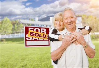Happy affectionate senior couple hugging in front of sold real estate sign and house