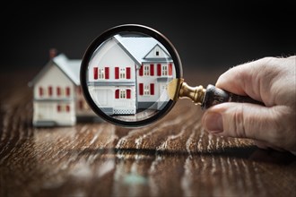 Hand holding magnifying glass up to model home on reflective wooden surface