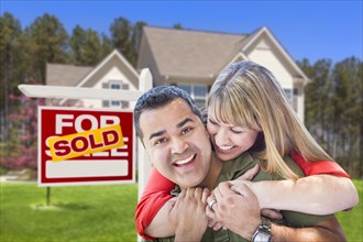 Happy hugging mixed-race couple in front of sold home for sale real estate sign and house