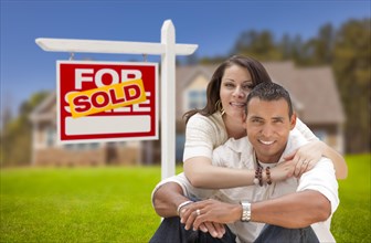 Young happy hispanic young couple in front of their new home and sold for sale real estate sign