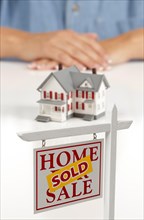 Womans folded hands behind model house and sold home for sale real estate sign in front on white surface
