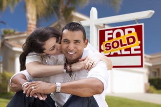 Young happy hispanic young couple in front of their new home and sold for sale real estate sign