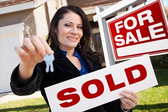 Happy attractive hispanic woman holding sold real estate sign and keys in front for sale real estate sign and house