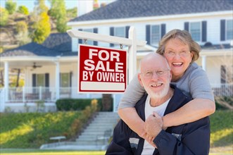 Senior adult couple in front of home for sale real estate sign and beautiful house