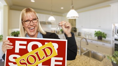 Happy young woman holding sold for sale real estate sign and keys inside beautiful custom kitchen
