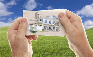 Male hands holding stack of paper with house drawing over empty grass field and sky