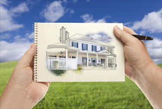 Male hands holding pen and pad of paper with house drawing over empty grass field and sky