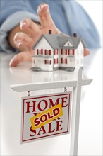Sold real estate sign in front of womans hand reaching for model house on a white surface