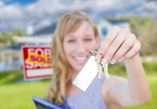 Woman holding new house keys with blank card in front of sold real estate sign and home