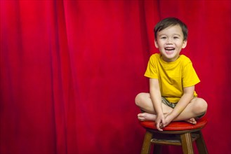 Laughing mixed-race boy sitting on stool in front of red curtain