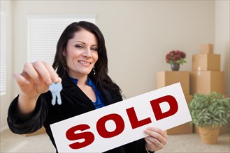 Happy hispanic female real estate agent with sold sign and keys in room with moving boxes