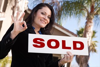 Happy attractive hispanic woman holding sold sign in front of house