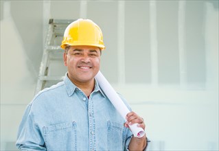 Hispanic male contractor with blueprint plans wearing hard hat in front of drywall and ladder