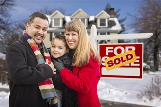 Warmly dressed young mixed-race family in front of sold home for sale real estate sign and house with snow on the ground