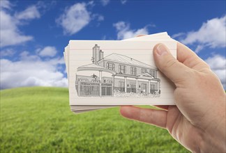 Male hand holding stack of paper with house drawing over empty grass field and sky
