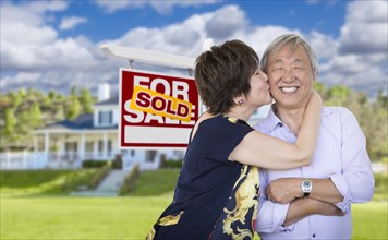 Attractive affectionate senior chinese couple in front of beautiful house and sold for sale real estate sign