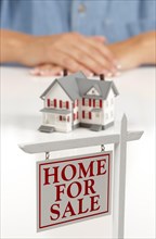 Womans folded hands behind model house and home for sale real estate sign in front on white surface