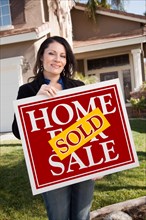Happy attractive hispanic woman holding red sold home for sale sign in front of house
