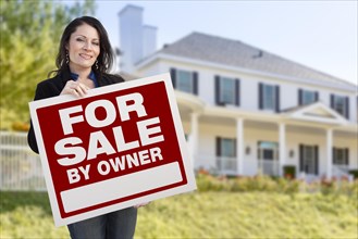 Smiling hispanic female holding for sale by owner sign in front of beautiful house