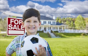 Playful young boy holding soccer ball in front of house and for sale real estate sign