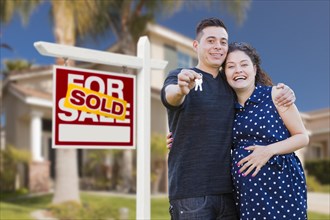 Young happy pregnant hispanic young couple with house keys in front of their new home and sold for sale real estate sign