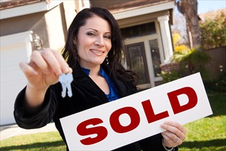 Happy attractive hispanic woman holding keys and sold sign in front of house