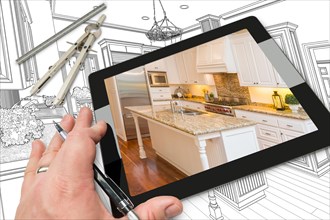 Hand of architect on computer tablet showing photo of kitchen drawing behind with compass and ruler