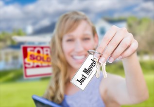 Woman holding new house keys with just moved card in front of sold real estate sign and home