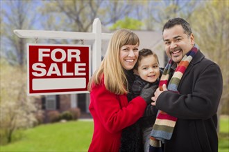 Warmly dressed young mixed-race family in front of home for sale real estate sign and house