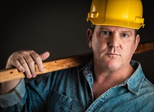 Serious contractor in hard hat holding plank of wood with dramatic lighting