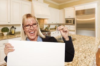 Happy young woman holding blank sign and keys inside beautiful custom kitchen