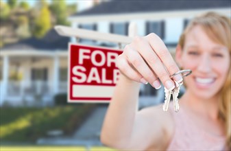 Excited woman holding house keys and for sale real estate sign in front of nice new home